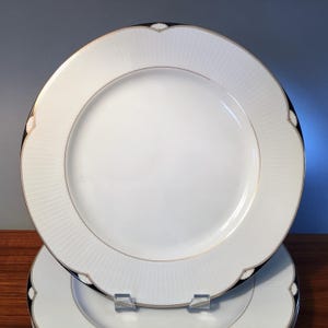 May include: Two white ceramic dinner plates with a decorative design. The plates have a white center, a textured rim with vertical lines, and a scalloped edge with black and gold accents. The plates are stacked on top of each other.