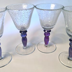 May include: Four clear wine glasses with a textured, bubble-like pattern. Each glass has a purple and blue beaded stem and a round base. The glasses are arranged on a white surface, showcasing their elegant design.