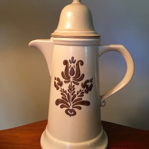 May include: Cream-colored ceramic coffee pot with a lid and handle. The pot features a brown floral design on the front. The pot is on a wooden surface.