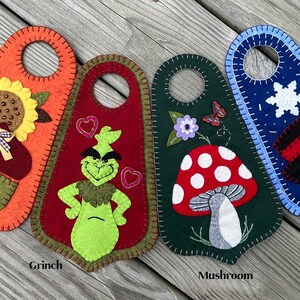 May include: Four felt door hangers with cutouts at the top. The orange one features an acorn, sunflower, and pumpkin. The red one shows a green character with hearts. The dark green one displays a mushroom and flower. The blue one depicts a snowman.