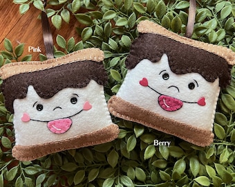 May include: Two felt s'mores ornaments, one pink and one red, with smiling faces and googly eyes. The ornaments are hanging from brown and pink ribbons.