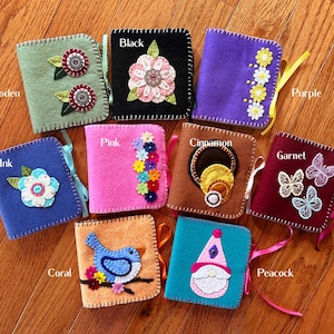 May include: A collection of handmade felt notebooks in various colors, including green, black, purple, blue, pink, brown, and coral. Each notebook features unique embroidered designs such as flowers, butterflies, and a bird. The notebooks are tied with colorful ribbons.
