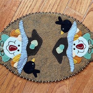 May include: Oval-shaped felt table mat with a whimsical design. Features two clown faces with colorful accents, black birds, and decorative elements. The mat is brown with a scalloped edge and small bead details.