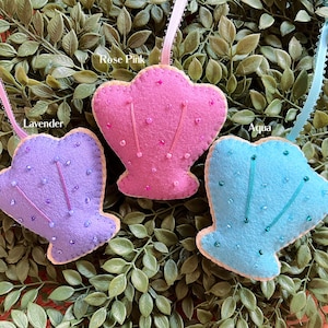 May include: Three felt seashell ornaments with beaded details. The seashells are in the colors lavender, rose pink, and aqua. The ornaments are labeled with their colors.