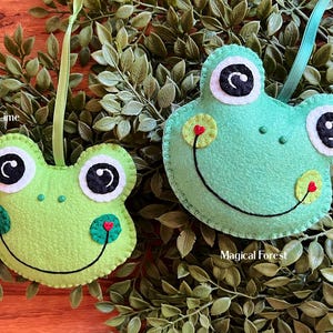 May include: Two felt frog ornaments, one green and one lime green, with black eyes and red hearts on their bellies. The green frog has the text "Magical Forest" below it, and the lime green frog has the text "Lime" above it.