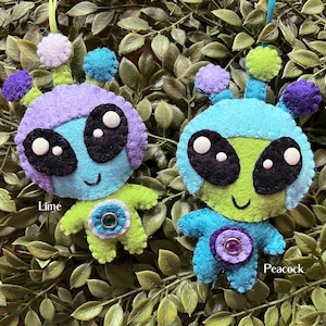 May include: Two felt alien ornaments, one green with a blue helmet and the other blue with a purple helmet. Both have black eyes and a smiling mouth. The green alien has a lime green body and the blue alien has a peacock blue body.  The text "Lime" is below the green alien and the text "Peacock" is below the blue alien.