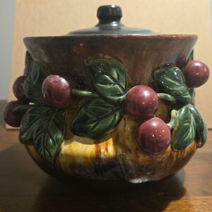 May include: A round ceramic jar with a dark brown lid, featuring a relief design of glossy red cherries and green leaves wrapped around the exterior. The base of the jar has a mottled brown and tan glaze with a smooth, reflective finish throughout.