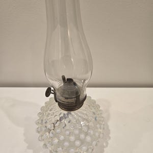 May include: A clear glass oil lamp featuring a round base with a raised hobnail texture and an iridescent finish. The lamp includes a metal burner assembly with a side adjustment knob and a tall, slender, clear glass chimney shade on top.