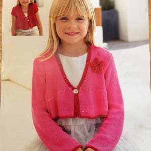May include: A pink knitted cardigan with a red trim and a red flower detail. The cardigan is shown on a young girl wearing a white dress. The pattern number is 2281 and the size is 2-13 years. The text "FLIRT DK" and "SIRDAR" are also visible.