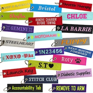 May include: A selection of embroidered keyrings in diverse colours, each bearing distinct text. The keyrings display phrases such as "Bristol," "Throttle Therapy," and "Diabetic Supplies." They have metal key rings for attachment.