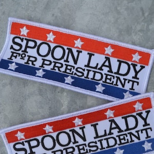 Può includere: Due toppe di tessuto bianco con stelle rosse, bianche e blu e il testo "Spoon Lady for President" in nero.