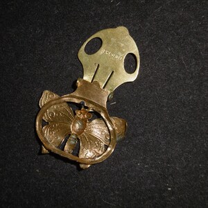 May include: Gold-toned metal butterfly-shaped clip with a decorative design. The clip has a spring mechanism and a small metal loop on the top. The back of the clip is engraved with the text "DEPOSE".