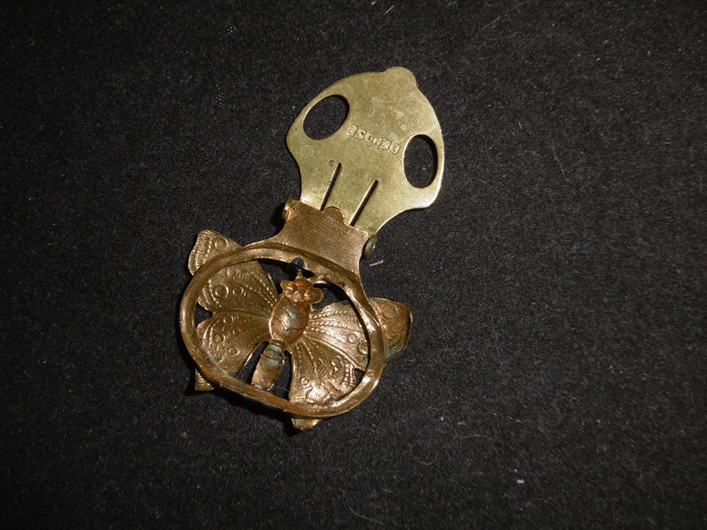 May include: Gold-toned metal butterfly-shaped clip with a decorative design. The clip has a spring mechanism and a small metal loop on the top. The back of the clip is engraved with the text "DEPOSE".