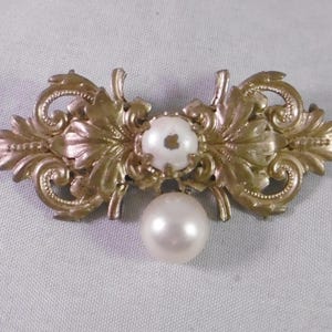 May include: A gold-toned brooch with an ornate, leaf-like design. The brooch features a central white pearl and a larger, dangling pearl. The design is symmetrical and detailed, with a vintage aesthetic.