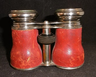 May include: A pair of vintage silver-toned opera glasses with red leather covering. The glasses have a center adjustment knob.