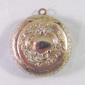 May include: A gold-toned locket with an ornate, engraved design. The locket has a hinged lid and a loop for attaching a chain.