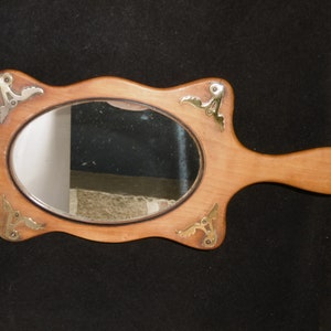 Puede incluir: Un espejo de mano de madera con un espejo de forma ovalada y un mango largo. El espejo tiene un marco decorativo con detalles en latón.