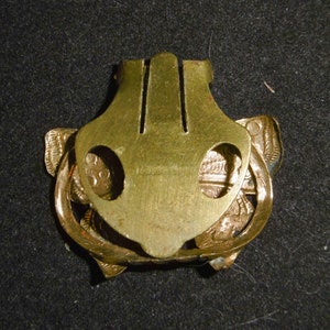 May include: A gold-toned metal clasp with a decorative design. The clasp has two holes on either side and a central section with a raised design.