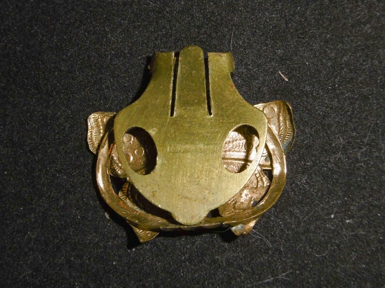 May include: A gold-toned metal clasp with a decorative design. The clasp has two holes on either side and a central section with a raised design.