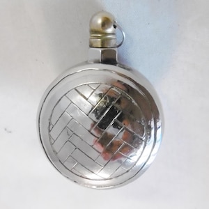 Peut inclure: Une fiole en métal argenté avec un motif tissé sur le devant. La fiole a un petit couvercle rond avec un bouton sur le dessus.