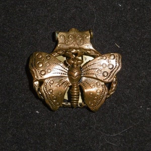 May include: A gold-toned metal butterfly brooch with intricate details. The butterfly has a detailed body and wings with a textured surface.