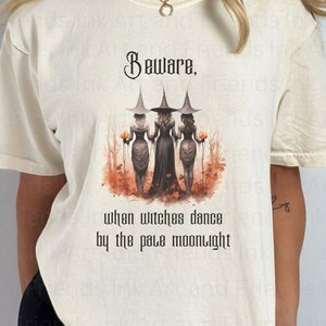 May include: A white t-shirt with a graphic of three witches dancing under a full moon. The text "Beware. When witches dance by the pale moonlight" is printed below the image.