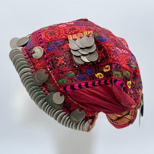以下が含まれることがあります： 銀貨の装飾が施された赤い刺繍の帽子。帽子は、さまざまな色とテクスチャの細かい模様が特徴です。帽子は白いマネキンの頭に展示されており、その形とデザインを際立たせています。