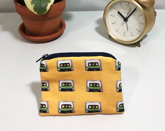 May include: A yellow zippered pouch with a repeating pattern of cassette tapes. The pouch is on a white surface next to a gold alarm clock.