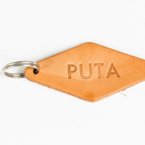 May include: A light brown leather keychain in a diamond shape with the word "PUTA" embossed on it. The keychain has a silver metal split ring attached for keys. The background is white.