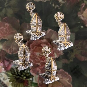 May include: Four silver and gold-toned metal charms shaped like a small building with a pointed roof and a small orb on top. The charms have intricate details and are set against a background of pink and green roses.