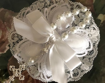 May include: White lace and satin flower with a silver rosary and cross charm. The rosary has white beads and a silver chain.