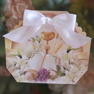 May include: A white favour box with a scalloped edge and a white satin ribbon bow. The box features a religious image of a chalice, bible, and lilies with the word "IHS" on the chalice.