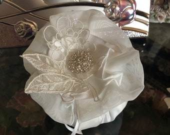 Könnte beinhalten: Ein weißes Satin-Ringkissen mit einem zarten Blumenmuster aus Spitze und einem glitzernden Strassstein in der Mitte.