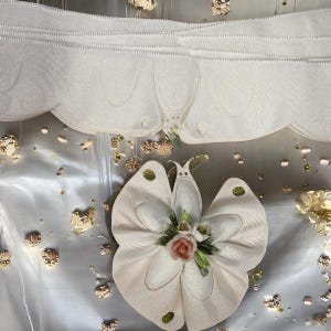 May include: A decorative butterfly made of white ribbon with a pink rose and green leaves in the center. The butterfly is surrounded by gold-colored confetti and a white scalloped ribbon.