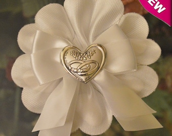 Könnte beinhalten: Weiße Satinblume mit einem silbernen Herz-Anhänger mit zwei ineinander verschlungenen Ringen und einem floralen Design. Der Anhänger ist an einer weißen Satin Schleife befestigt.