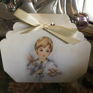 May include: A white favour box with a scalloped edge and a white satin ribbon. The box features a watercolour illustration of a young boy holding a bouquet of flowers and a small bird.