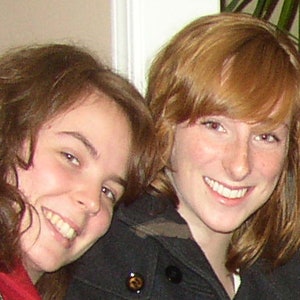 May include: Two young women with long brown and red hair smiling at the camera. The woman with red hair is wearing a black jacket and the woman with brown hair is wearing a red scarf.