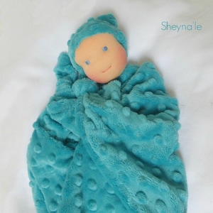 May include: A teal-colored baby doll swaddled in a matching plush blanket. The doll has a light peach face with rosy cheeks, blue eyes, and a small smile. The blanket and hat have a textured, dotted pattern. The text "Sheyna'le" is in the upper right corner.