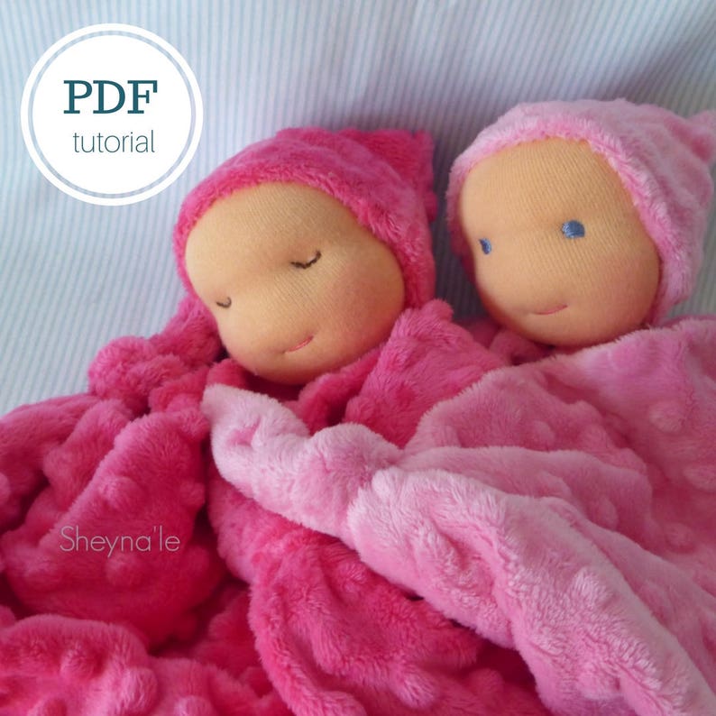 May include: Two pink fabric dolls with closed eyes and pink hats are lying on a pink and white textured blanket. The dolls are wearing pink and white blankets. The text "Sheyna'le" is visible in the lower left corner of the image.