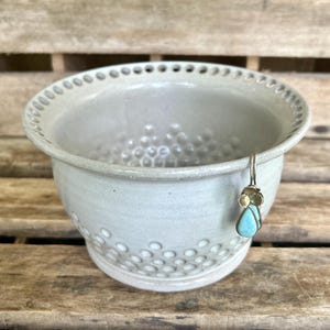 May include: A light gray ceramic bowl with a textured surface and a decorative rim. A gold-colored earring with a turquoise stone hangs from the bowl's edge. The bowl is on a wooden surface.