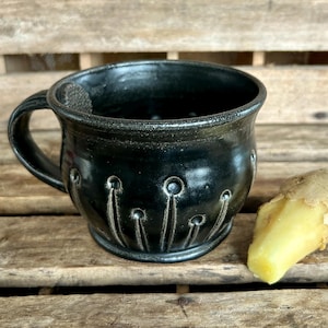 May include: A handmade black ceramic mug with a handle and a decorative pattern of vertical lines with circles. A piece of fresh ginger is next to the mug. The mug is on a wooden surface.