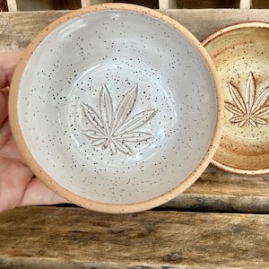 May include: Two ceramic bowls with a speckled glaze and a leaf design. The bowl on the left is white with a gray leaf design. The bowl on the right is yellow with a brown leaf design.