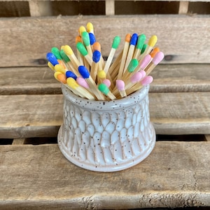 May include: A white ceramic match holder with a textured surface, filled with colorful matches. The matches have blue, green, yellow, and pink tips.