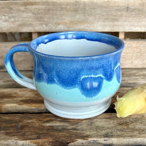 May include: A handmade ceramic mug with a blue and turquoise glaze. The mug has a rounded shape with a handle and sits on a white base. A piece of fresh ginger is next to the mug, suggesting a beverage.