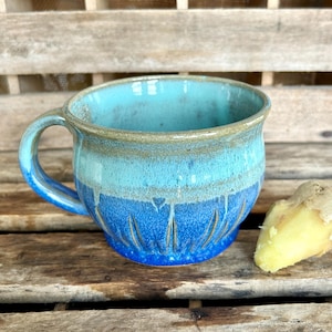 May include: A handmade ceramic mug with a blue and turquoise glaze. The mug has a rounded shape with a handle and decorative carved lines. A piece of fresh ginger sits beside the mug on a wooden surface.