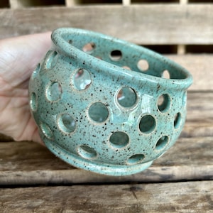 May include: A speckled, turquoise ceramic bowl with numerous circular cutouts. The bowl has a rounded shape with a slightly flared rim and a small, flat base. The surface has a speckled texture with dark brown spots.
