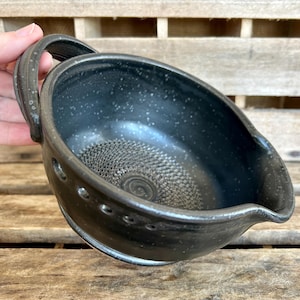 May include: A handmade, dark gray ceramic gravy boat with a handle and spout. The interior features a textured, spiral design. The exterior has small, white speckles and a series of small holes near the rim. The boat is held by a hand.