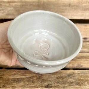May include: A handmade, off-white ceramic bowl with a skull and crossbones design embossed on the interior. The bowl has a slightly angled rim and a series of small, decorative holes around the exterior.