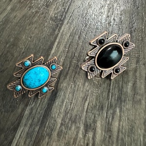 Op de afbeelding: Twee decoratieve metalen ringen met een geometrisch ontwerp. Een ring heeft een turquoise steen, terwijl de andere een zwarte steen heeft.