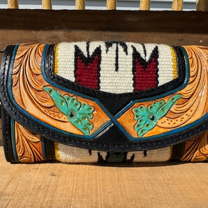 May include: A brown leather wallet with tooled floral designs and turquoise accents. The front flap features a woven panel with red and white stripes and the text "MTM".